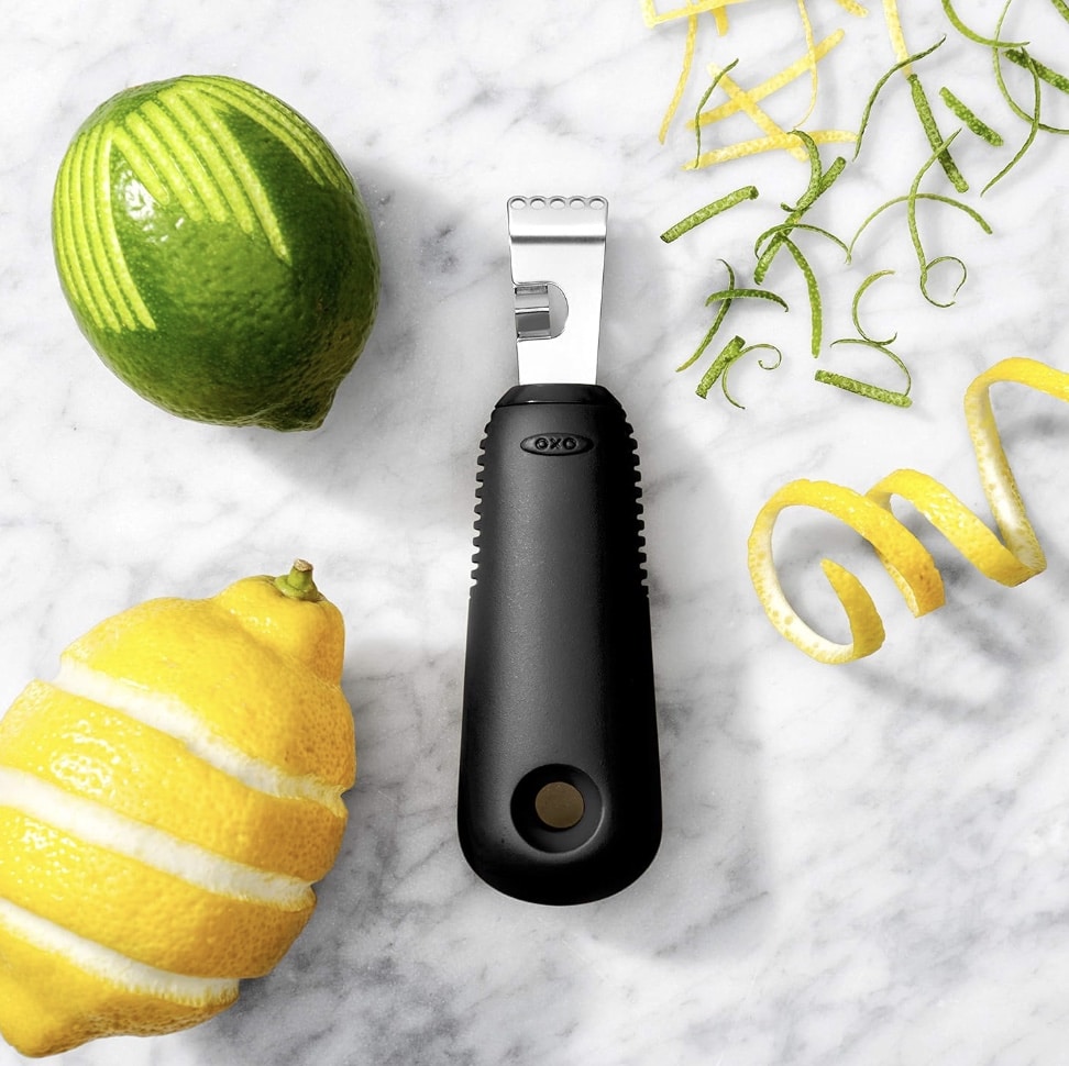 A citrus zester with a black handle sits on a marble surface, surrounded by a partially peeled lemon and lime, along with curly strips of zest—just the right tools for crafting that perfect healthy lemon loaf recipe.